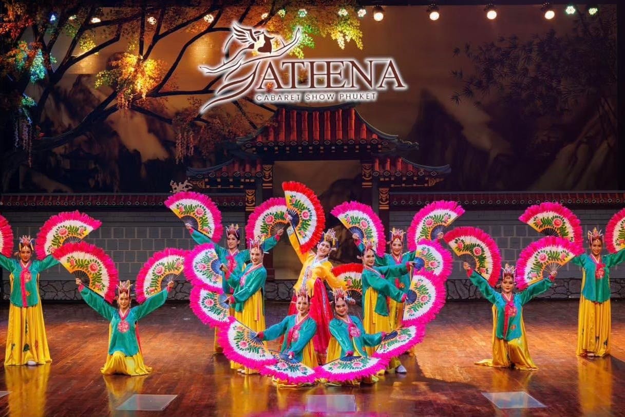 Des artistes en tenue traditionnelle colorée tiennent de grands éventails roses et verts sur une scène à l'arrière-plan décoratif lors du spectacle Athena Cabaret.