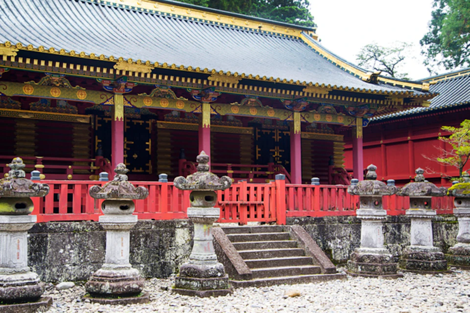 A traditional Japanese temple with ornate red and gold details, stone lanterns, and a tiled roof, surrounded by trees.