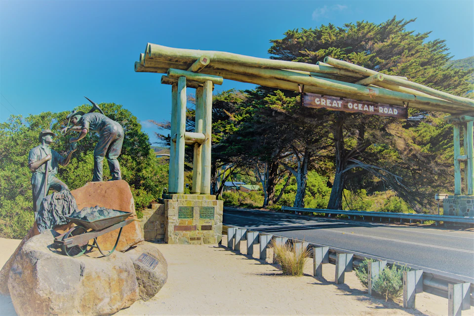 A stone archway labeled "Great Ocean Road," next to a sculpture of a worker with a wheelbarrow, surrounded by trees.