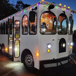 A white trolley bus adorned with colorful string lights is parked at dusk. The interior is lit, and the door is open.