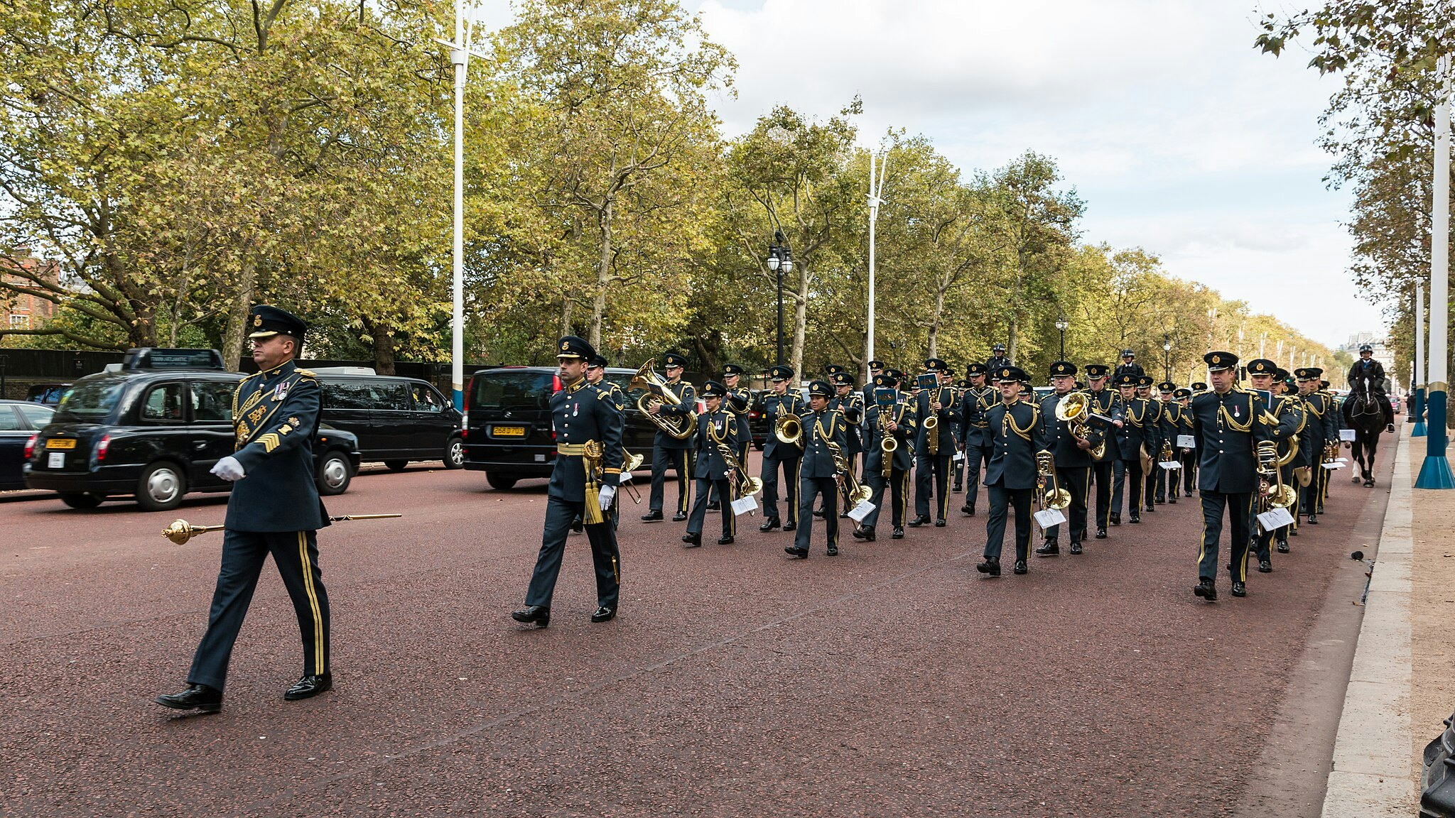 Uma banda marcial em uniformes militares formais desfila numa rua arborizada com táxis pretos ao fundo.