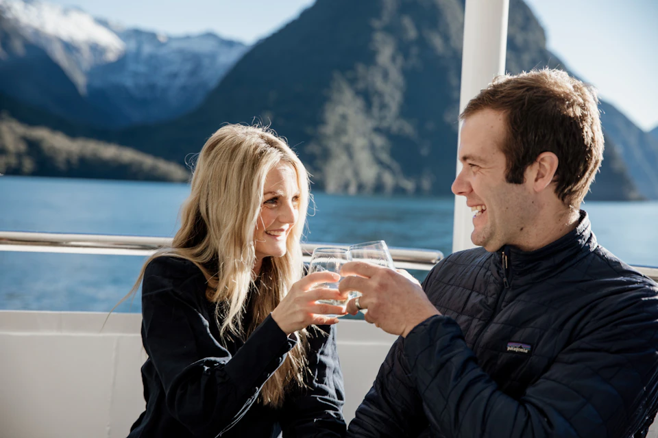 A man and a woman smiling while clinking glasses on a boat with a mountainous landscape in the background.