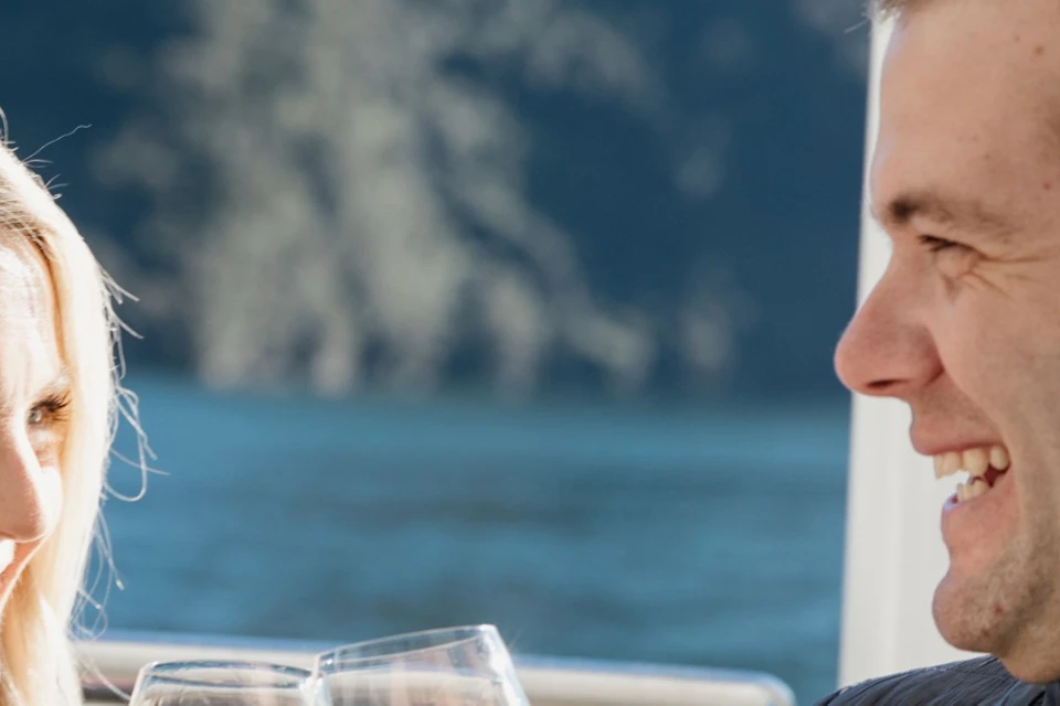 A man and a woman smiling while clinking glasses on a boat with a mountainous landscape in the background.