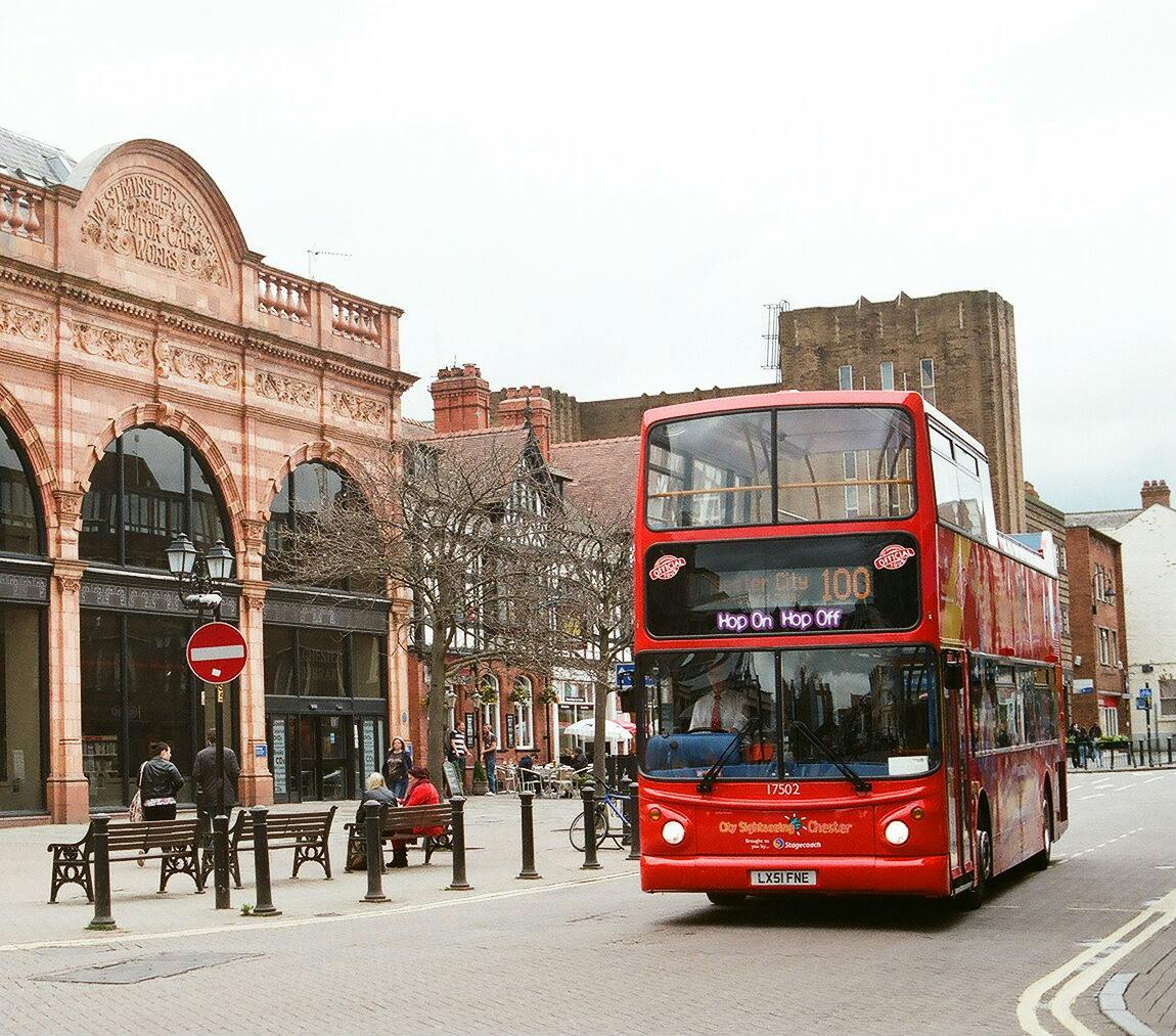 这辆红色双层巴士标有「随上随下」（ Hop On Hop Off ）字样，位于一条有历史建筑和行人的街道上。