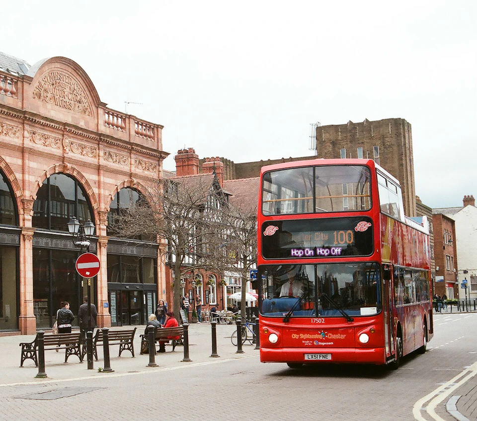 A red double-decker bus labeled "Hop On Hop Off" on a street with historic buildings and pedestrians.