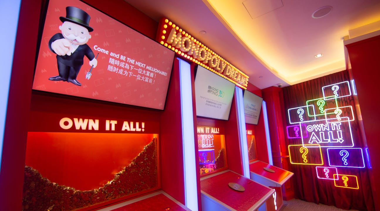 Une exposition interactive haute en couleur sur le thème du Monopoly, avec des néons, des écrans et une pancarte sur laquelle on peut lire « Own It All ! » en gros caractères.