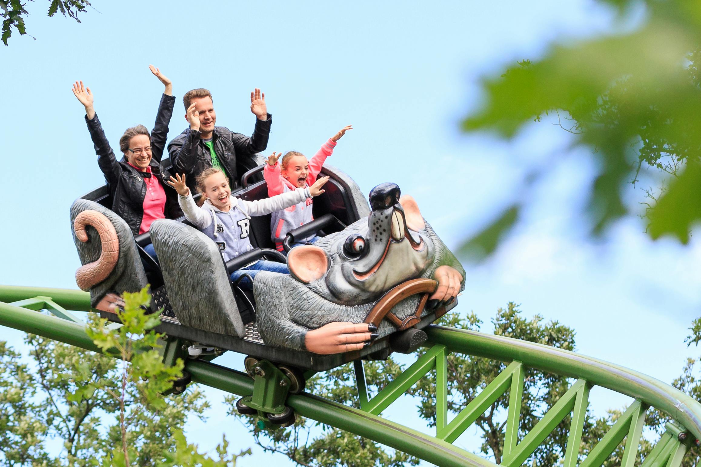 Quatre persones viatgen en una muntanya russa amb forma de ratolí, amb les mans alçades i les cares somrient, envoltades d'arbres i un cel clar.