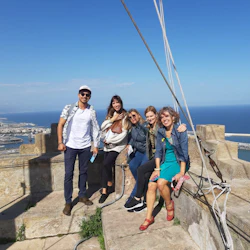 Fem personer poserar på en stenstruktur med en kuststad och blått hav i bakgrunden under en klar, solig himmel.