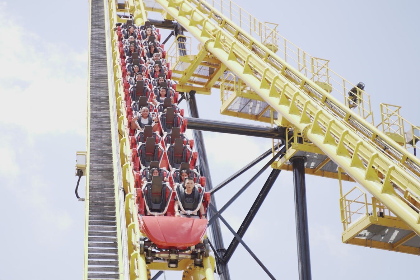 Za slunečného dne stoupá po strmé žluté dráze červené auto na horské dráze plné lidí.