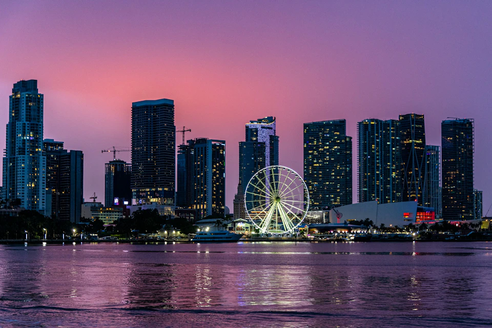 City skyline at dusk with illuminated Ferris wheel and reflections on a calm body of water.