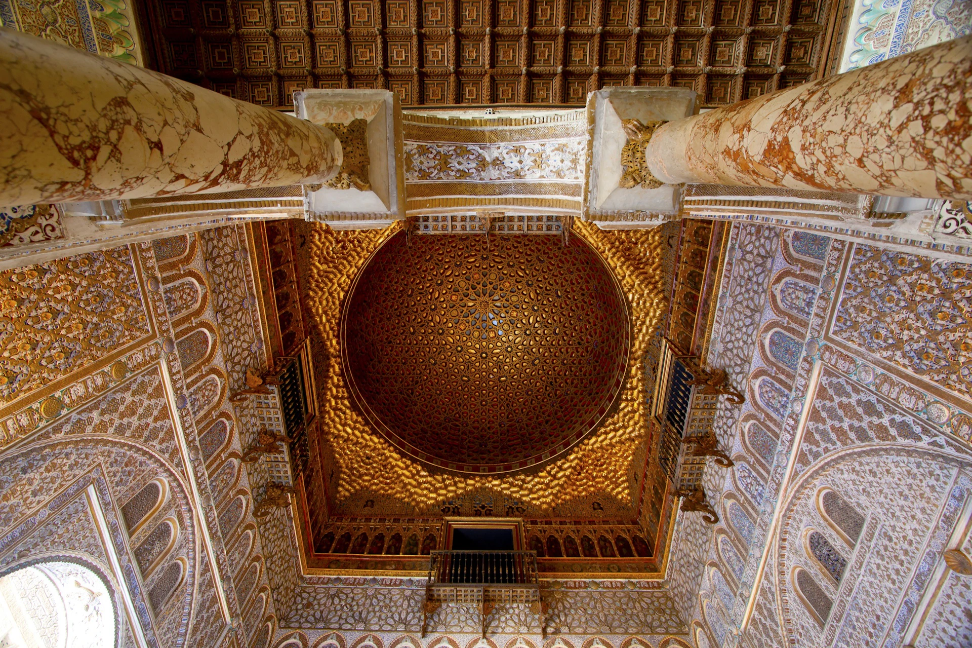A richly decorated ceiling with intricate patterns and a golden dome, surrounded by ornate walls and columns.
