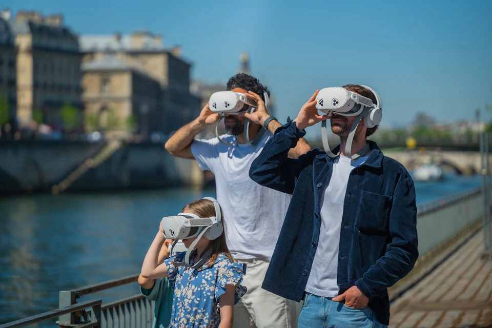 Three people, two adults and a child, wear VR headsets outdoors near a body of water and a historic building.