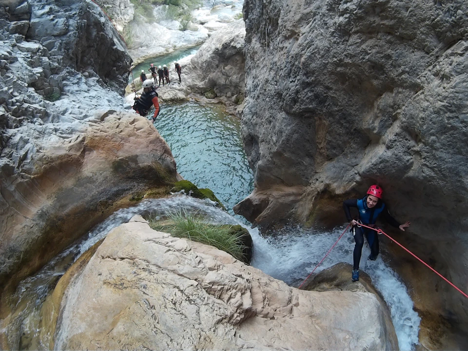 People wearing helmets and harnesses rappel down a rocky waterfall in a canyon with a stream below. Others stand in the stream.