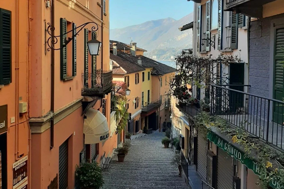 Narrow cobblestone street between colorful buildings with shutters, plants, lanterns, and a distant view of mountains under a clear sky.