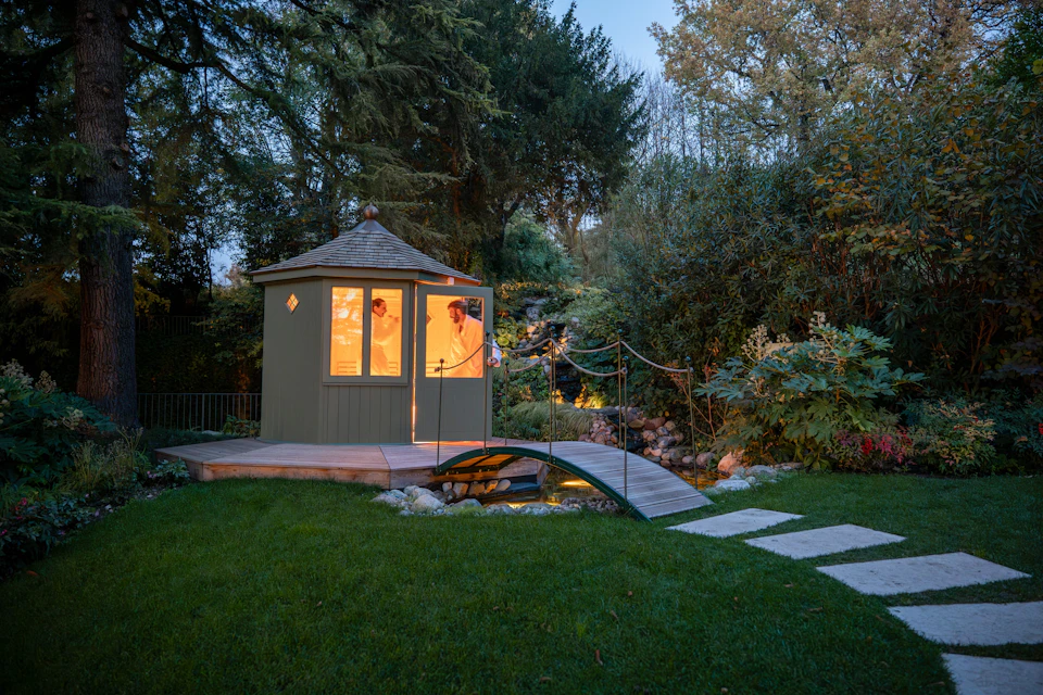 A small wooden hut with lit windows in a garden, surrounded by trees, a bridge, and stepping stones on a lawn at dusk.