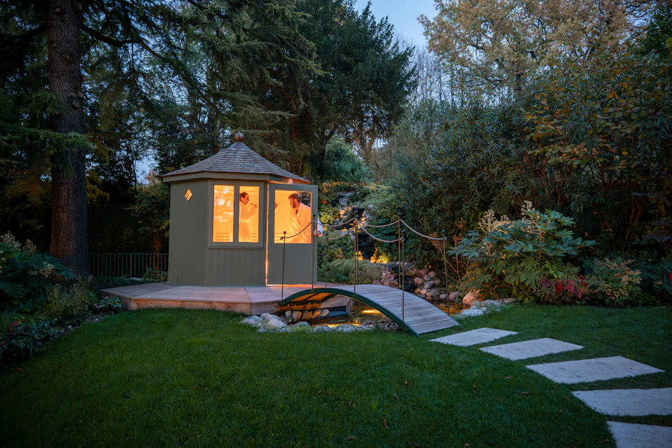 A small wooden hut with lit windows in a garden, surrounded by trees, a bridge, and stepping stones on a lawn at dusk.
