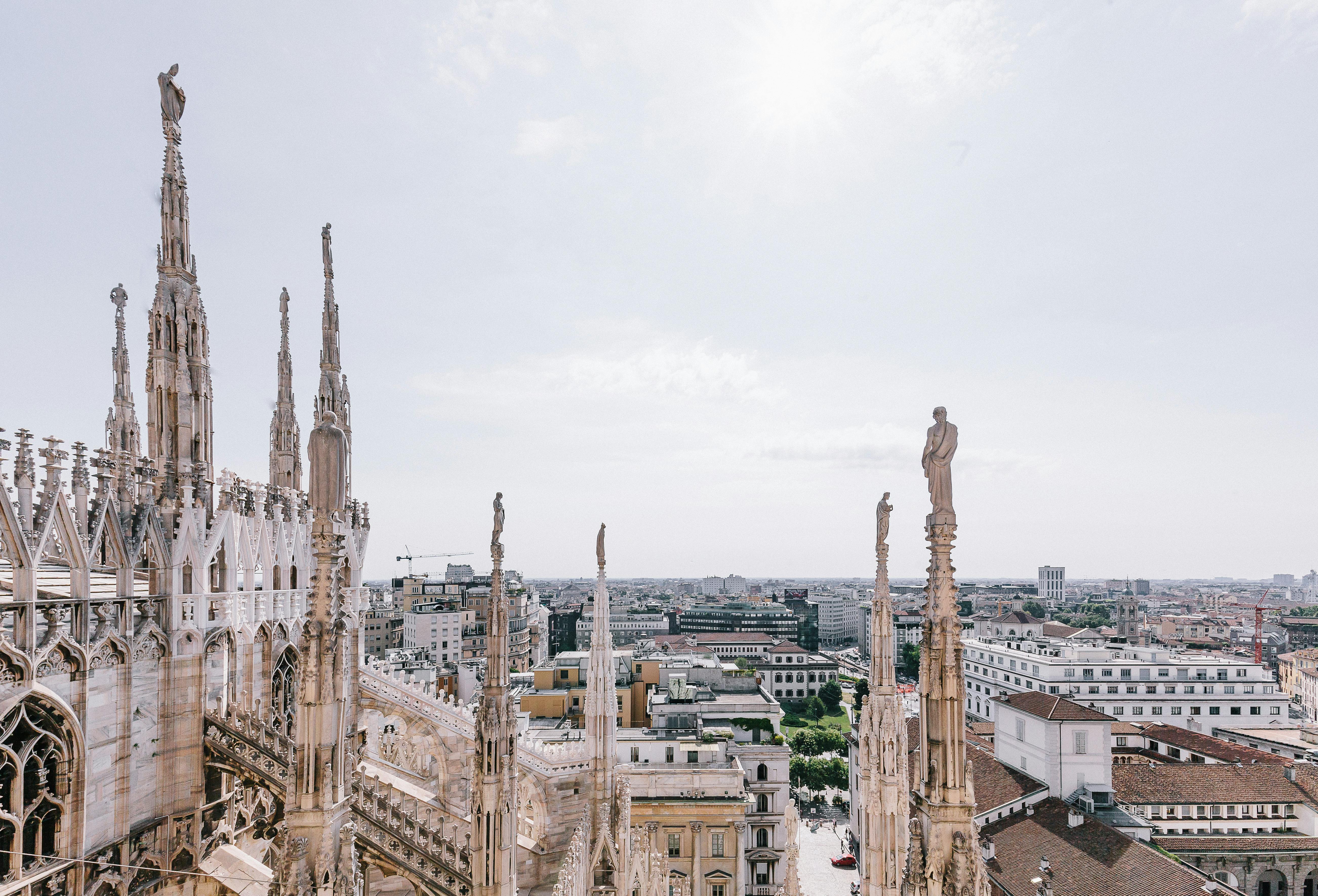Gothic spires and statues of a cathedral overlook a cityscape under a bright, clear sky.