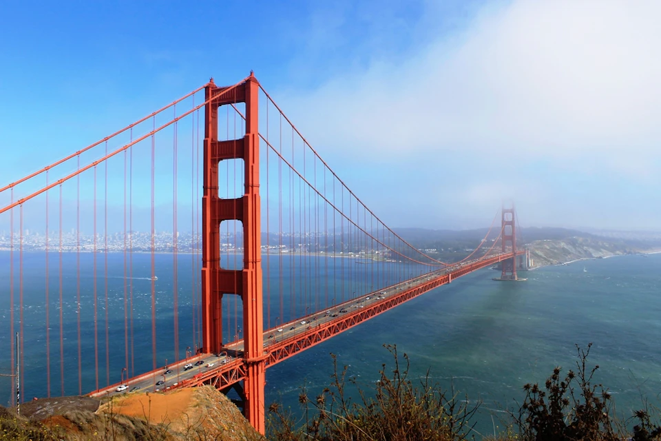 The image shows the Golden Gate Bridge spanning over water, connecting land areas under a clear blue sky with some distant fog.
