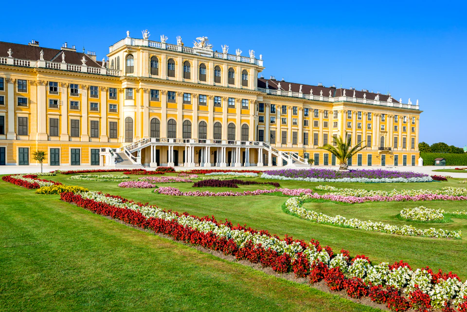 Yellow baroque palace with symmetrical gardens featuring colorful flower beds, trimmed grass, and a palm tree under a clear blue sky.