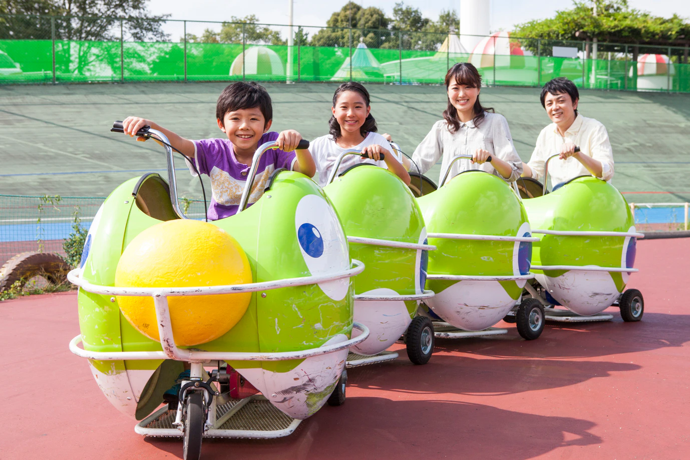 Children riding green and yellow pedal cars shaped like cartoon animals on a sunny day at an outdoor park.