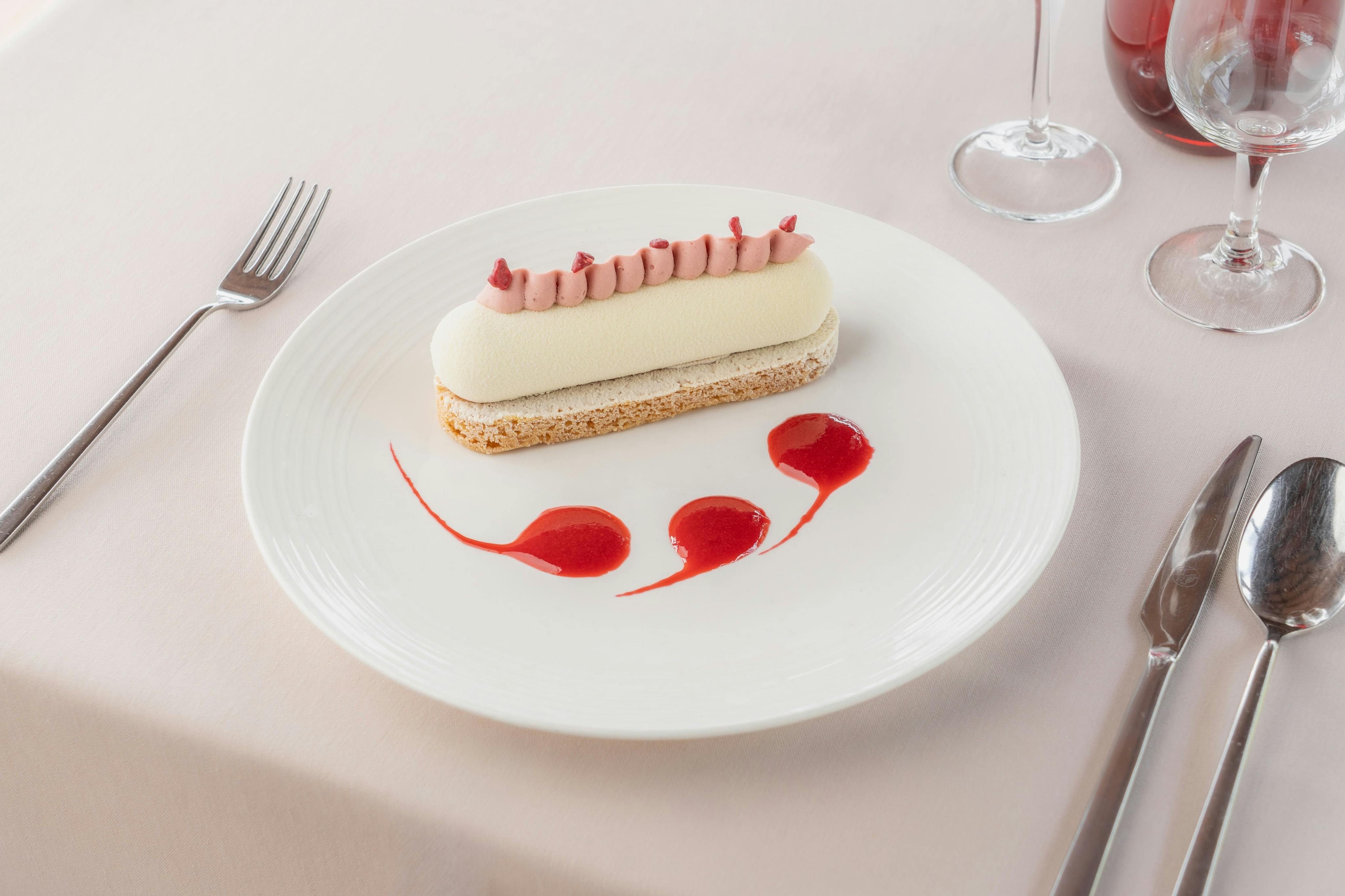 Un postre emplatat que consisteix en un tracte de mousse blanc amb decoració rosa, acompanyat de punts de salsa vermella, sobre un mantell blanc.