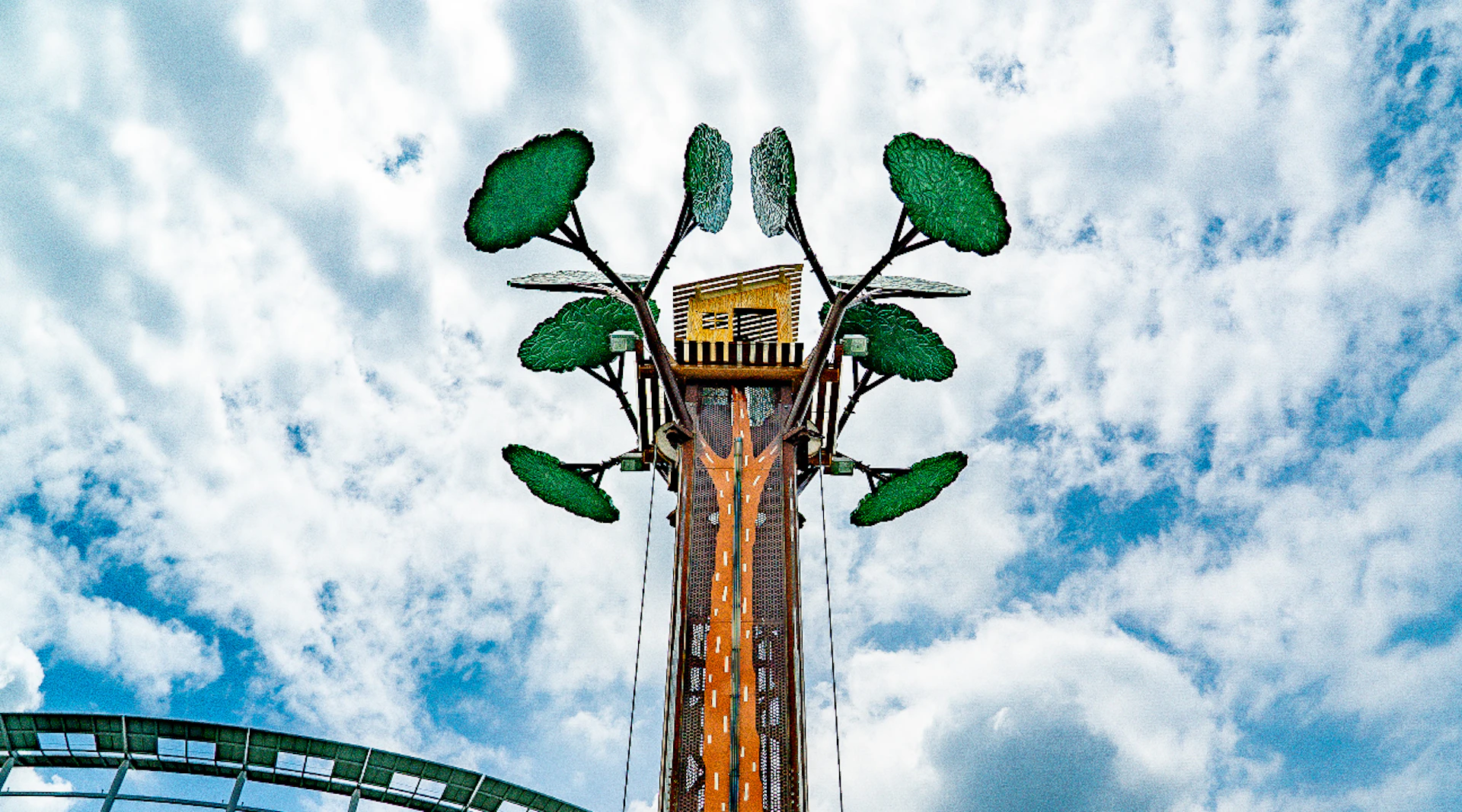 A tall amusement park ride designed like a tree with green foliage and a cabin at the top under a cloudy sky.
