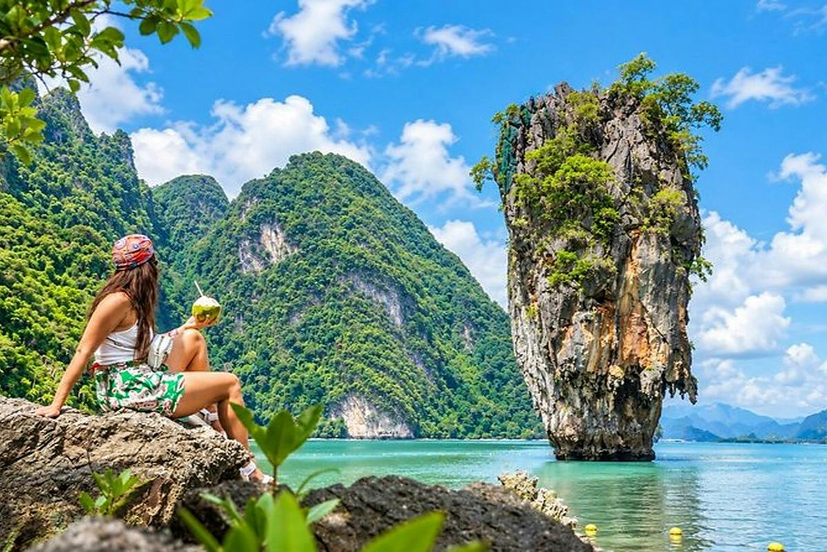 Two people sitting on rocks by a turquoise sea, with lush green islands and a tall rocky island in the background.
