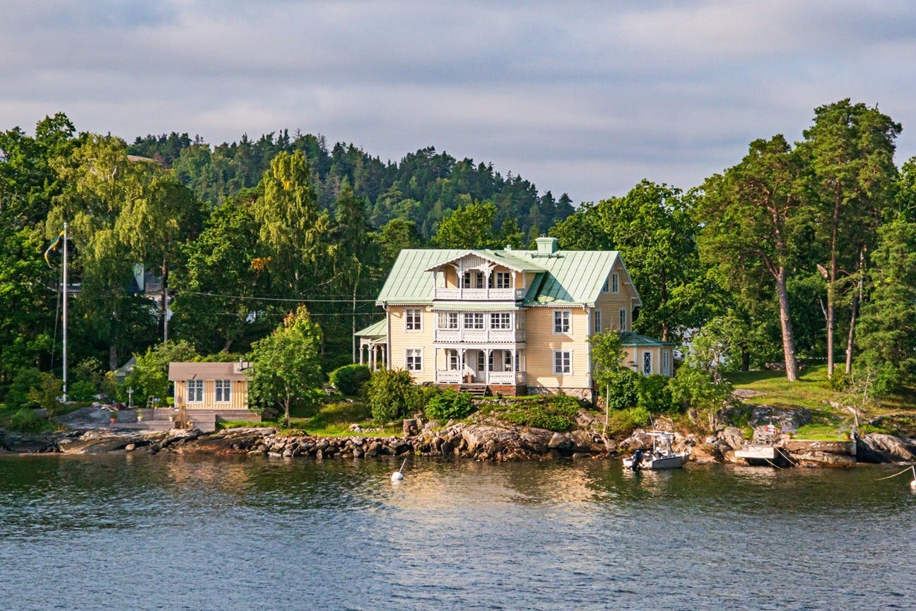 Casa groga amb teulada verda al costat de l'aigua, envoltada d'arbres. A mà dreta es pot veure un petit moll i un vaixell.