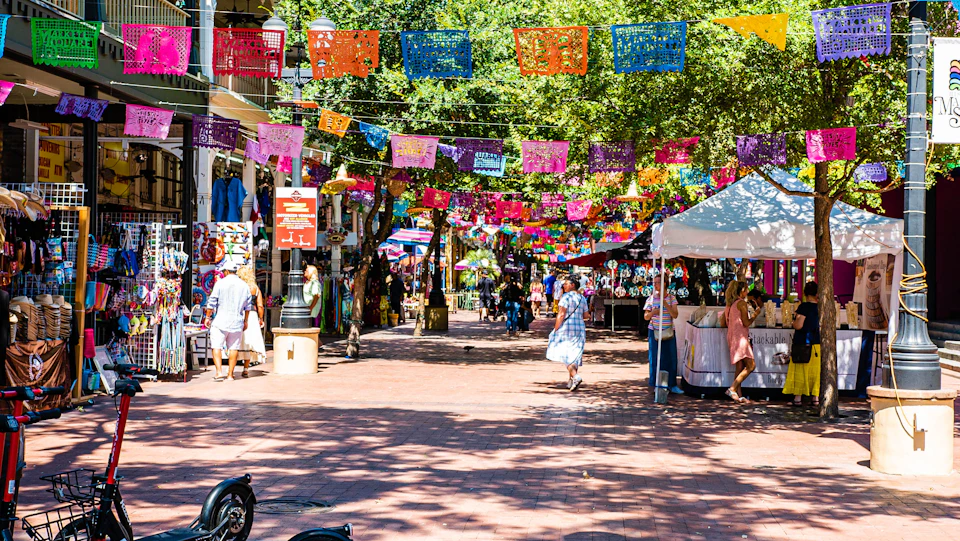 A vibrant outdoor market scene with colorful banners hanging, people shopping, and various stalls under the trees.