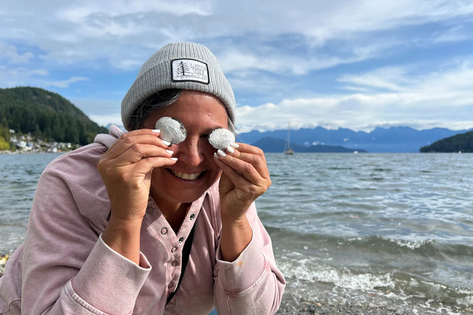 Person wearing a beanie and holding seashells over their eyes, smiling at a beach with mountains and sky in the background.