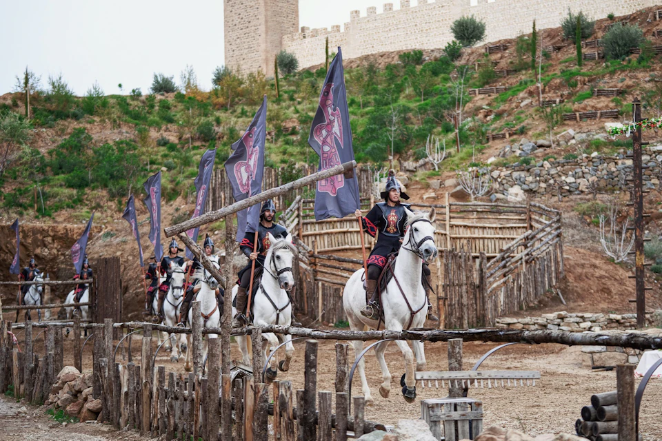 A group of riders on white horses carrying flags, dressed in medieval-style attire, ride past wooden structures near a castle.