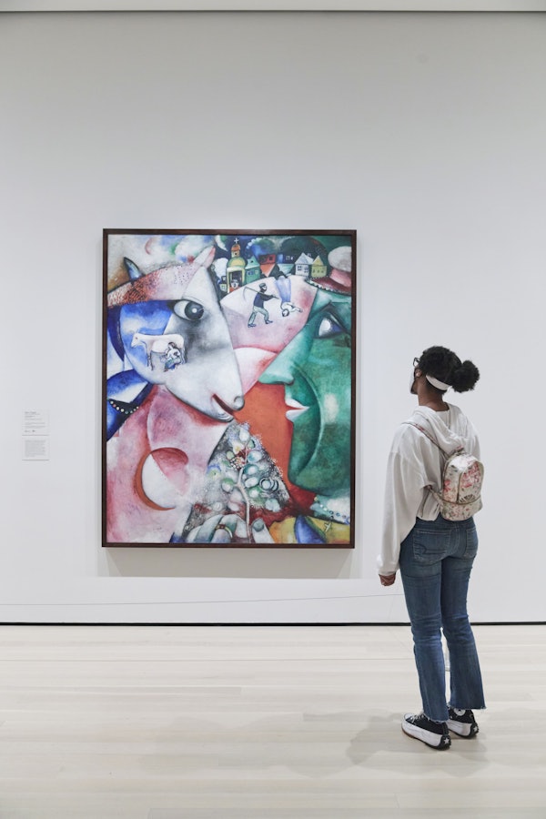 Person, der ser et farverigt abstrakt maleri i et kunstgalleri, iført en hvid hættetrøje og en rygsæk.