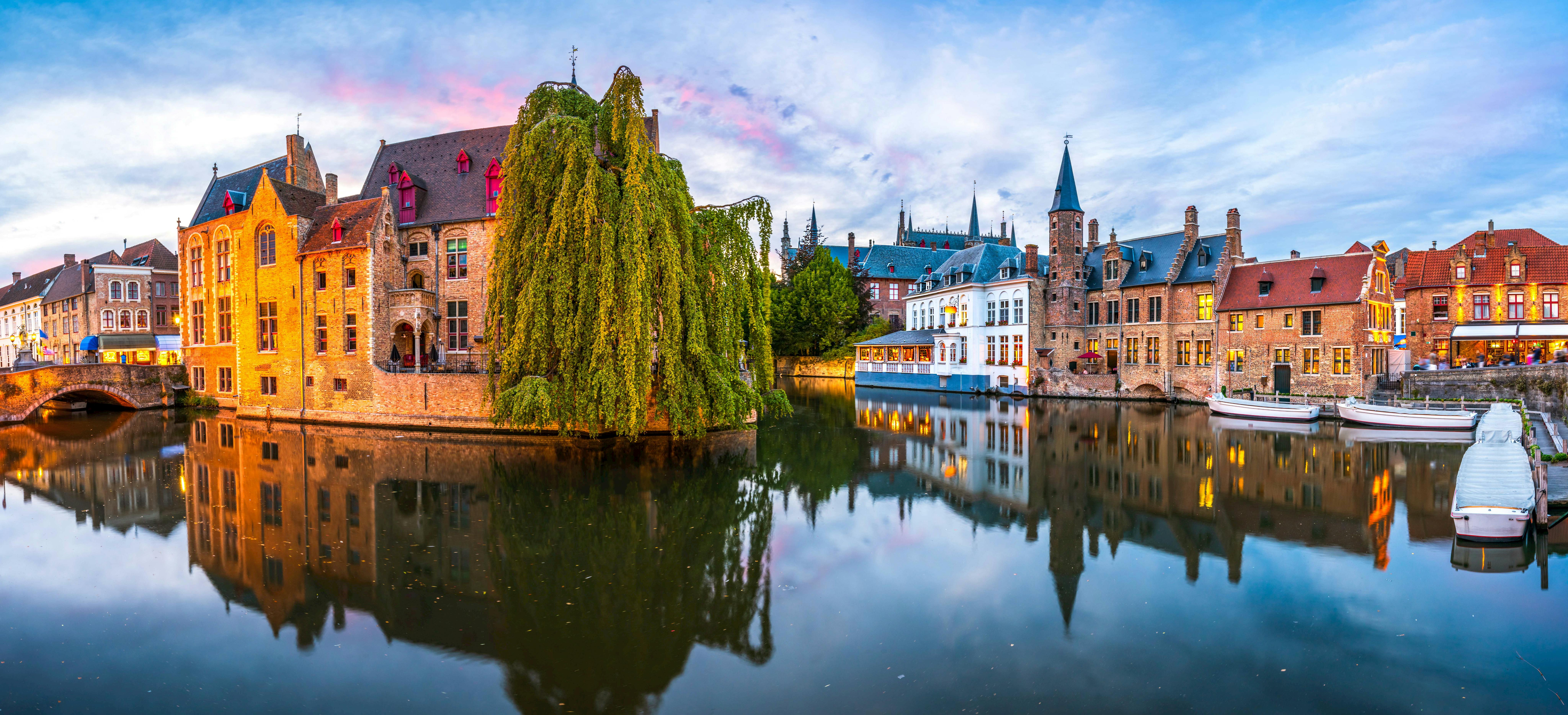 A scenic view of a canal with historic buildings and a large weeping willow tree reflecting in the water under a colorful sky.