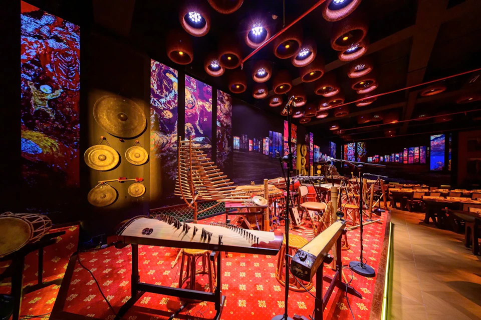 A room filled with traditional musical instruments, vibrant wall art, and patterned red carpet under illuminated circular ceiling lights.