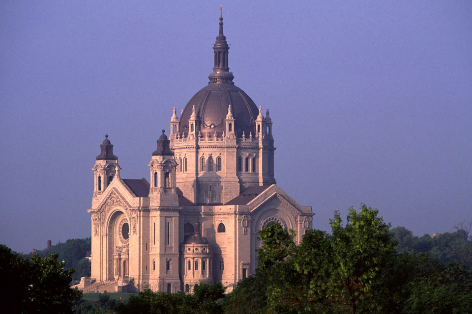 A grand cathedral with a large dome and spires, viewed from a distance, surrounded by trees under a clear sky.