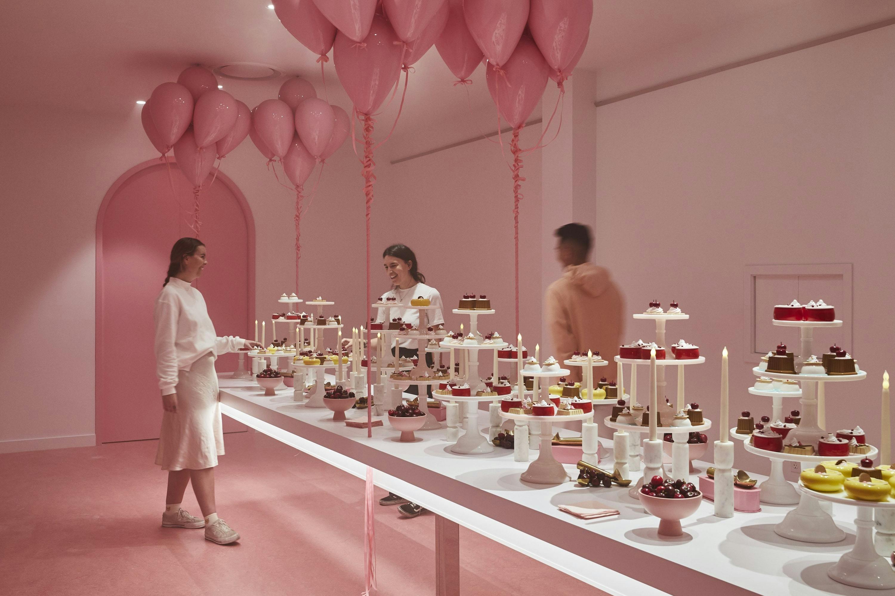 Tři lidé obdivují komplikovaný dezert na dlouhém stole s mnoha růžovými balónky v místnosti v pastelových barvách.