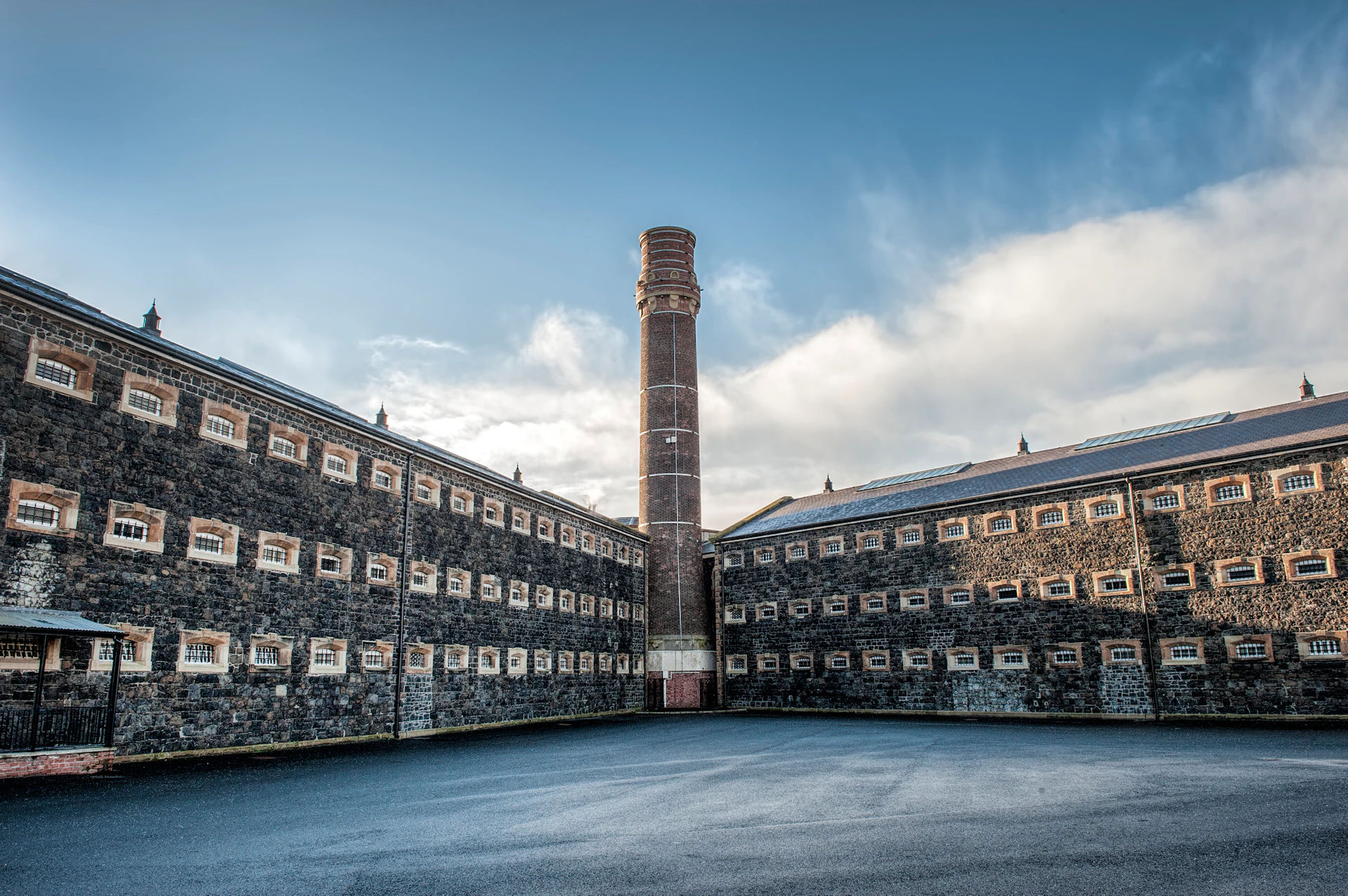 A large brick tower rises between two connected stone buildings under a blue sky with scattered clouds.