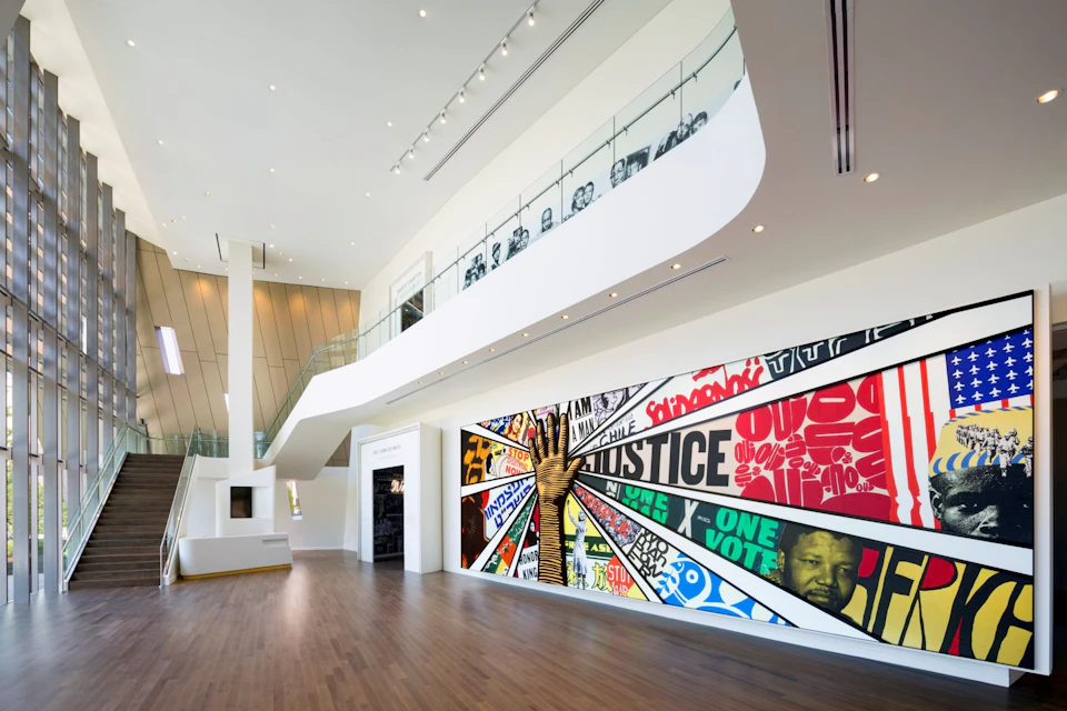 A modern interior space with a colorful mural displaying text and graphics on a wall, and a mezzanine with glass railings above.