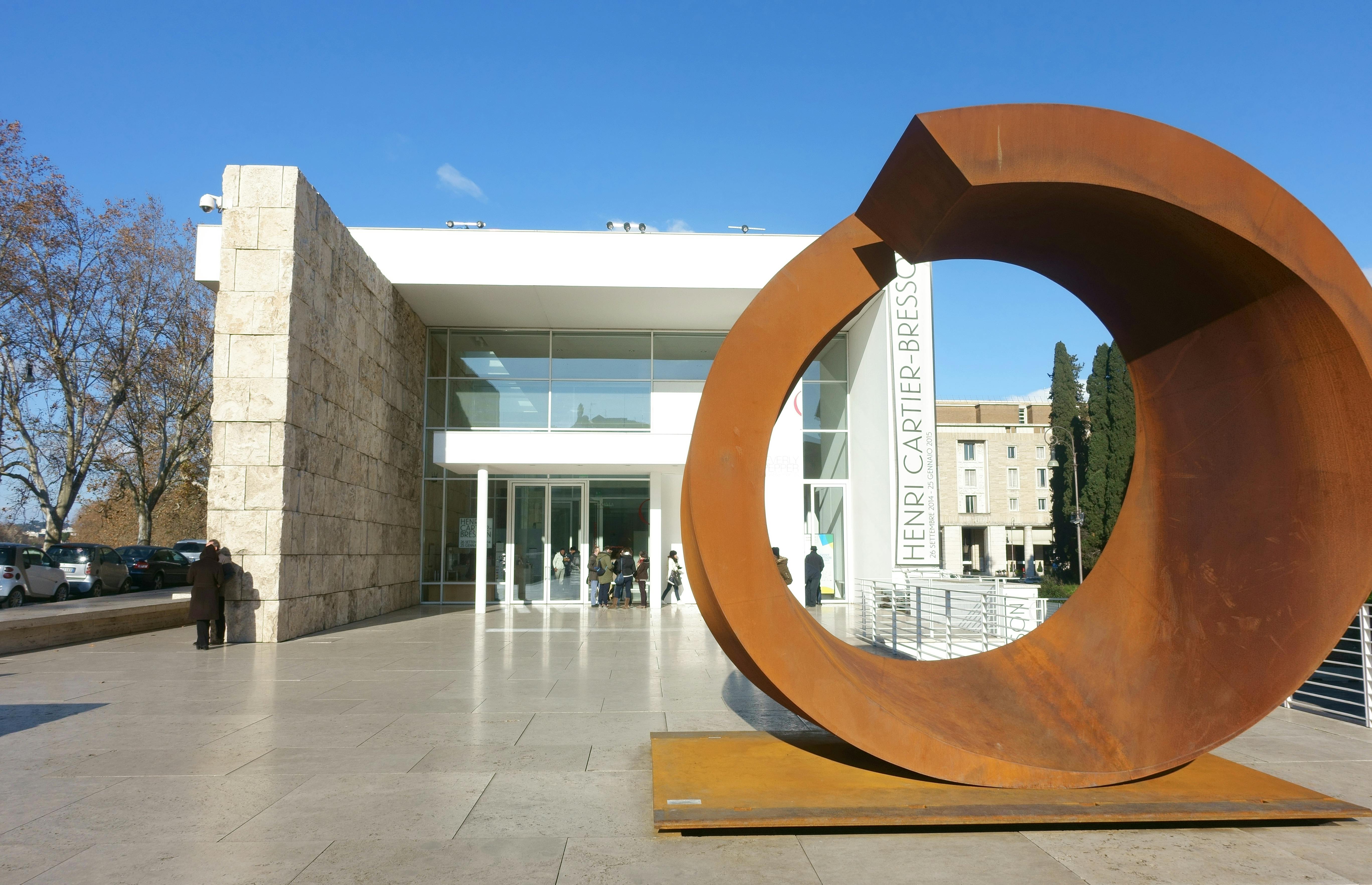 Large rusted metal sculpture in front of a modern white building with a sign that reads "Henri Cartier-Bresson." People are near the entrance.