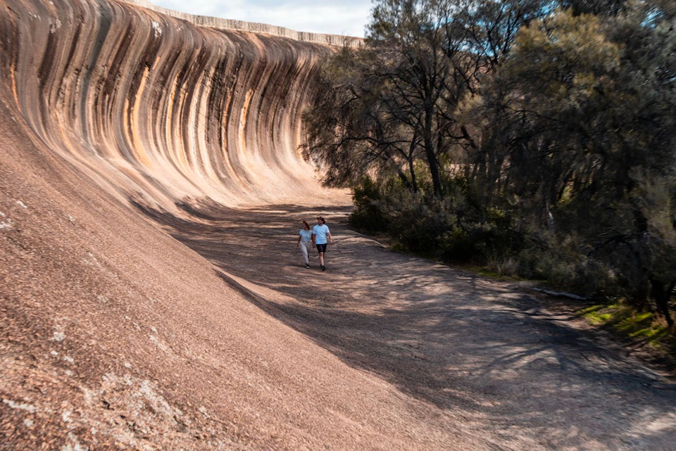 Two people walking along the curved rock face of Wave Rock, surrounded by sparse trees and sunlight filtering through.