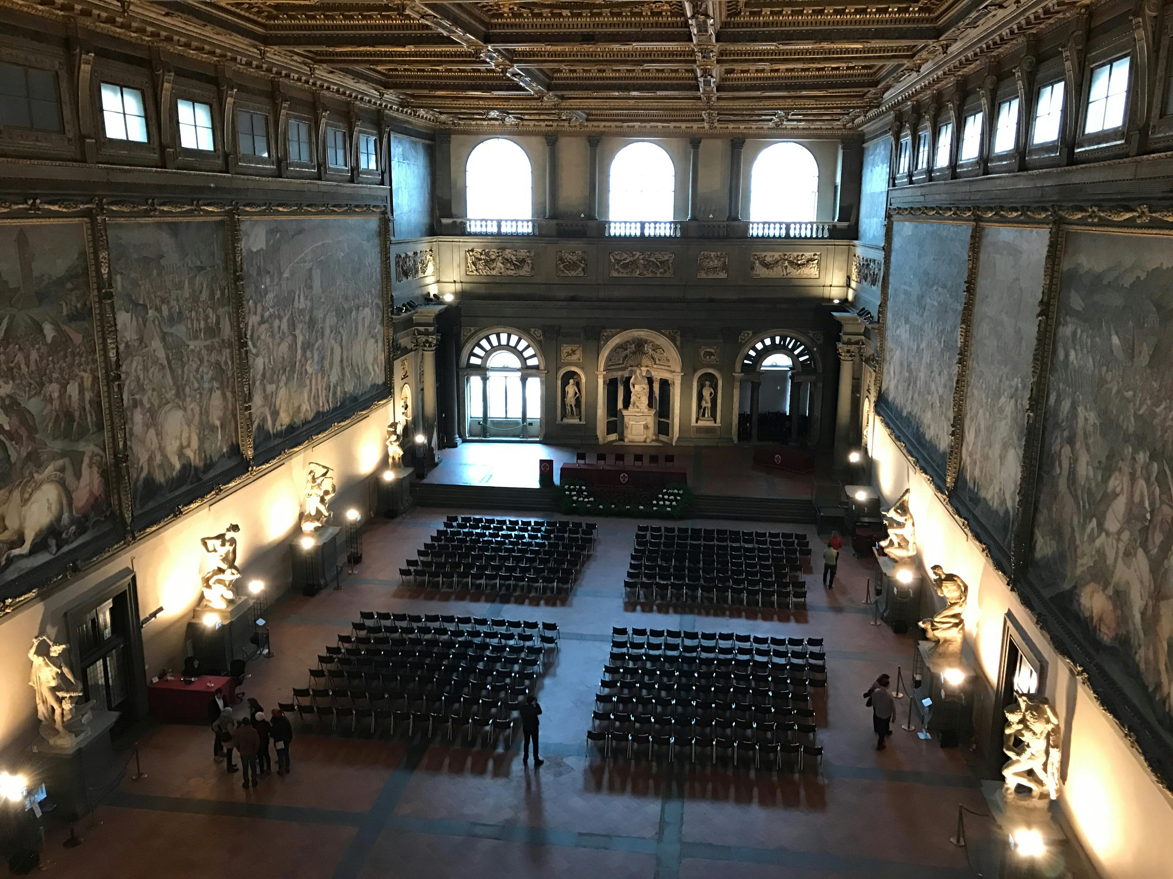 Grande sala con soffitto ornato, grandi murales e file di sedie disposte per un evento. Alcune persone sono in piedi e conversano.