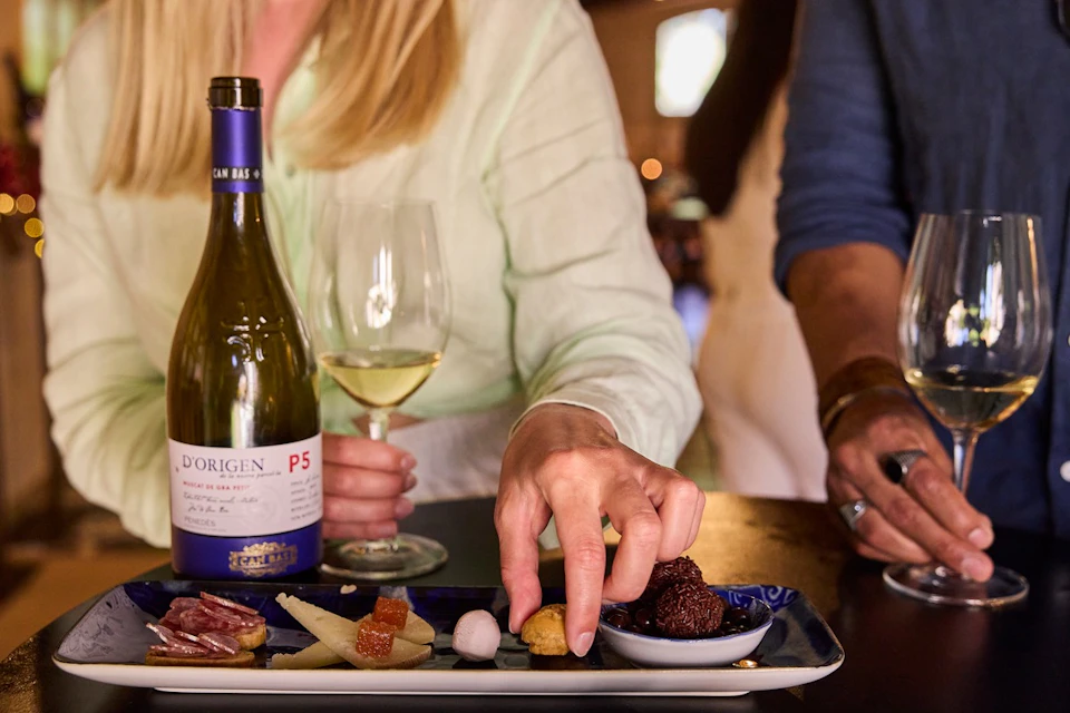 A person reaching for food on a plate with various items, next to a glass of white wine and a wine bottle on a table.