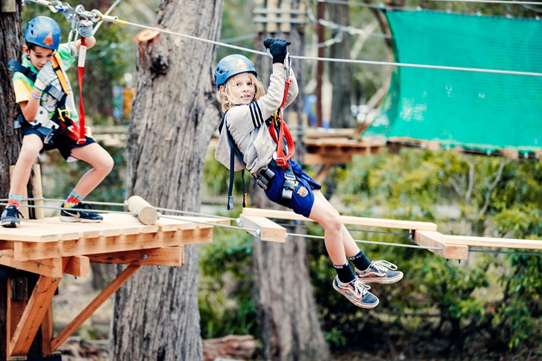 헬멧과 하네스를 착용한 두 명의 어린이가 나무 꼭대기 로프 코스를 탐색하고, 한 명은 플랫폼에서 균형을 이루고, 다른 한 명은 집라인을 착용합니다.