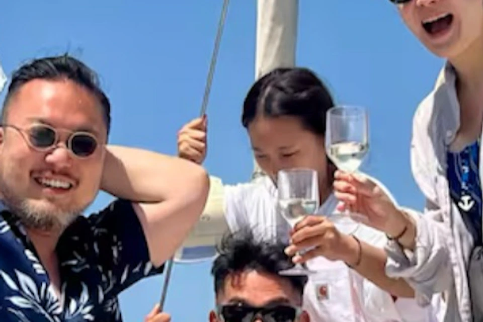 Four people on a sailboat, smiling and raising wine glasses, wearing casual summer attire and sunglasses against a clear, blue sky.