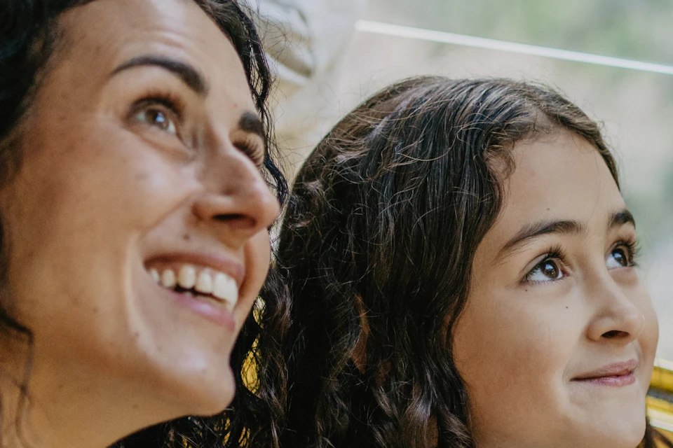 A woman and a young girl smile while looking out a window in a bright, natural setting.