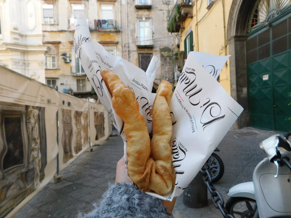 Two fried pastries wrapped in paper held by a hand, with old buildings and a street in the background.
