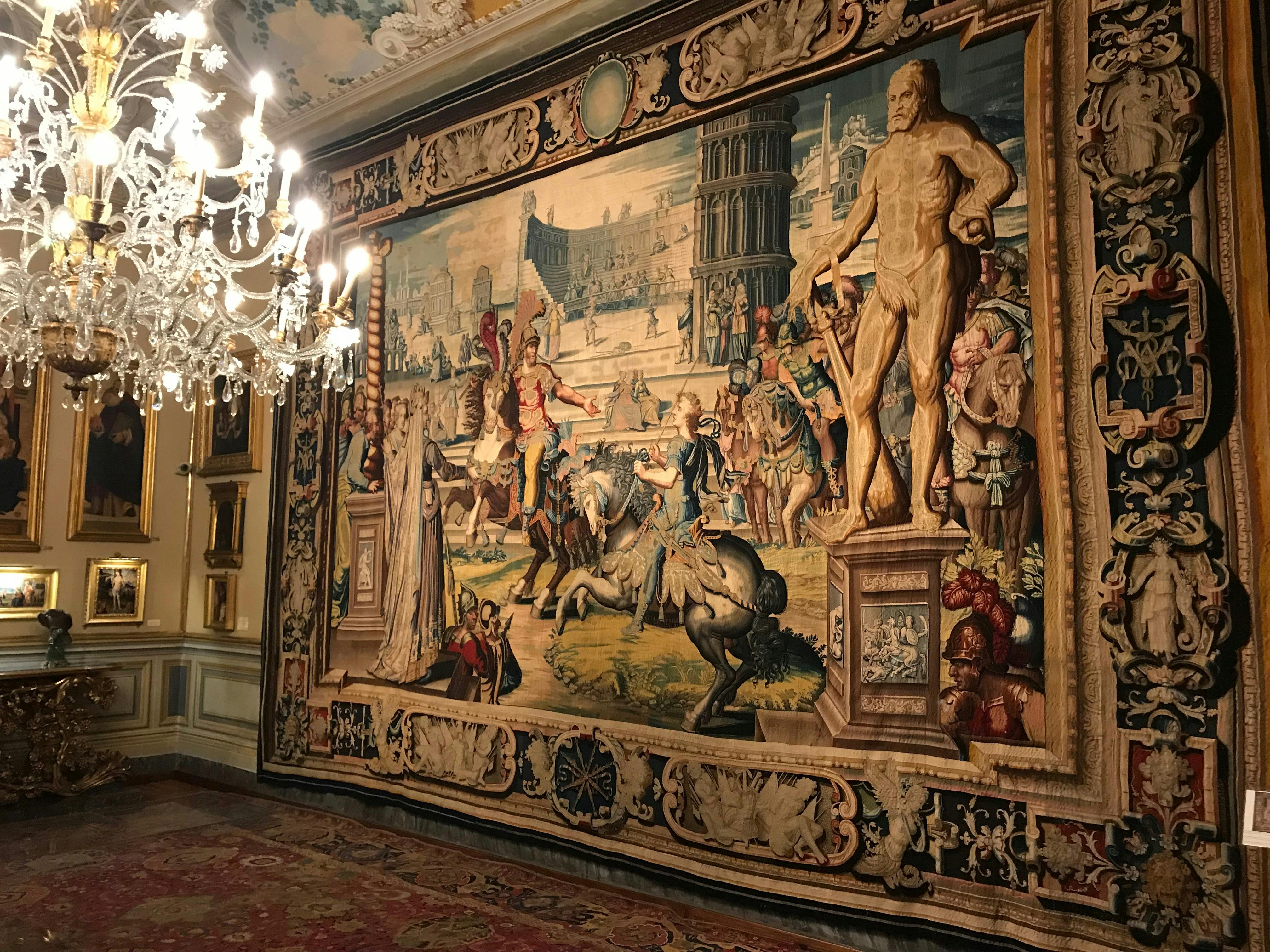 An ornate room with a large, intricate tapestry depicting historical scenes, a crystal chandelier, and a detailed ceiling.