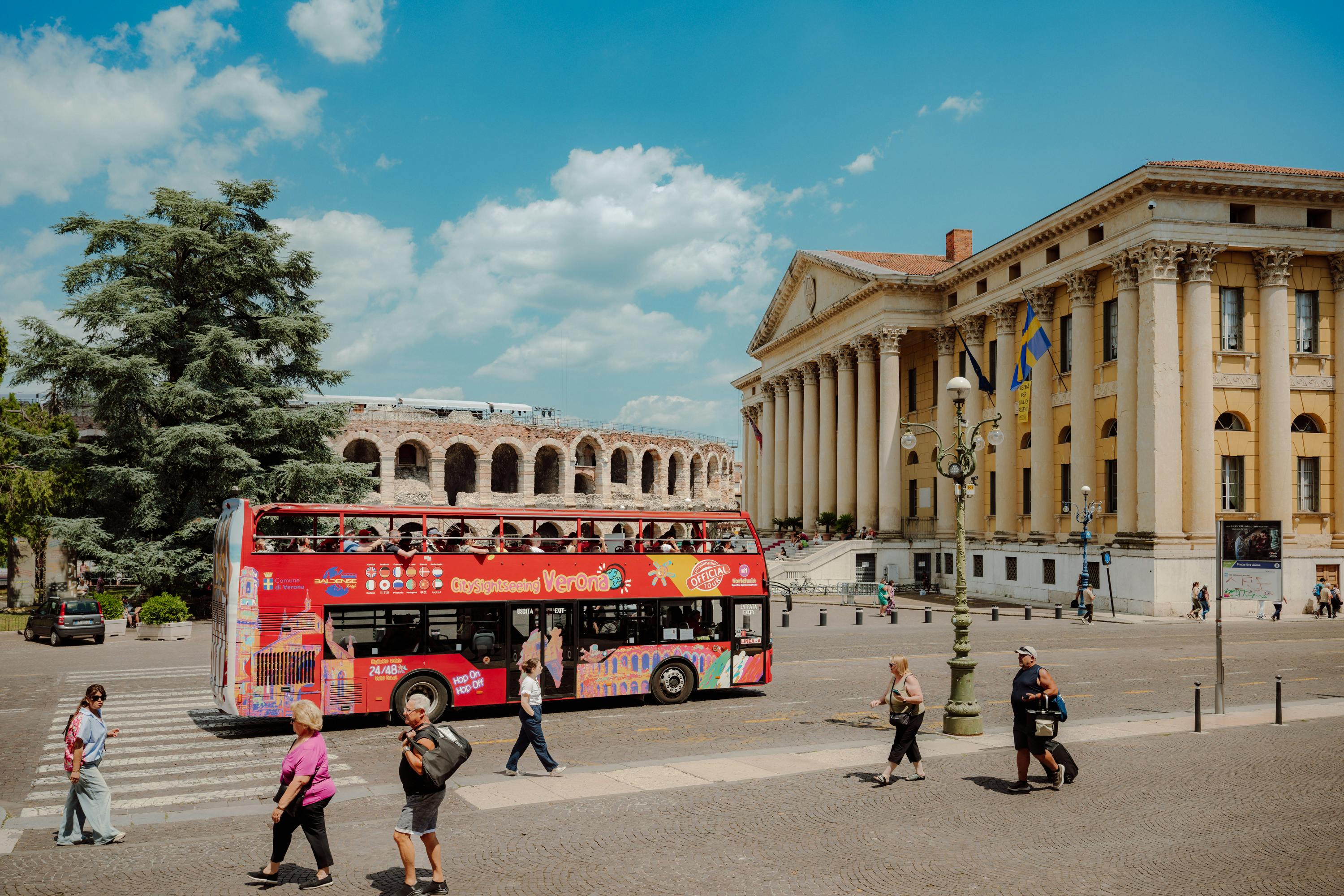 Ένα κόκκινο διώροφο τουριστικό λεωφορείο μπροστά από ένα νεοκλασικό κτίριο και αρχαία ερείπια σε μια ηλιόλουστη μέρα.