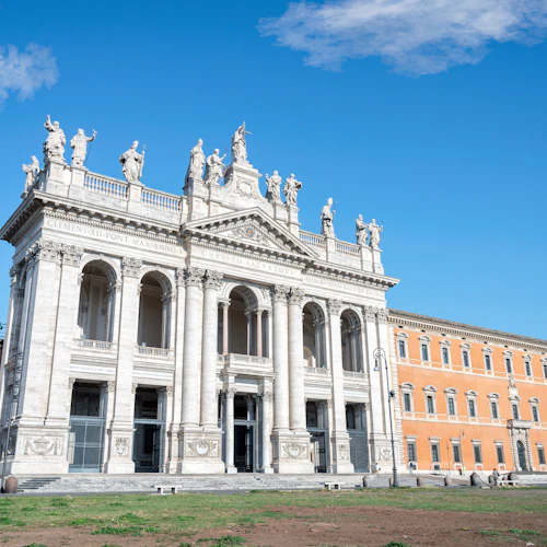 Historisk kyrkfasad med statyer, välvda fönster och pelare under en klarblå himmel, i anslutning till en kupol och orange byggnad.