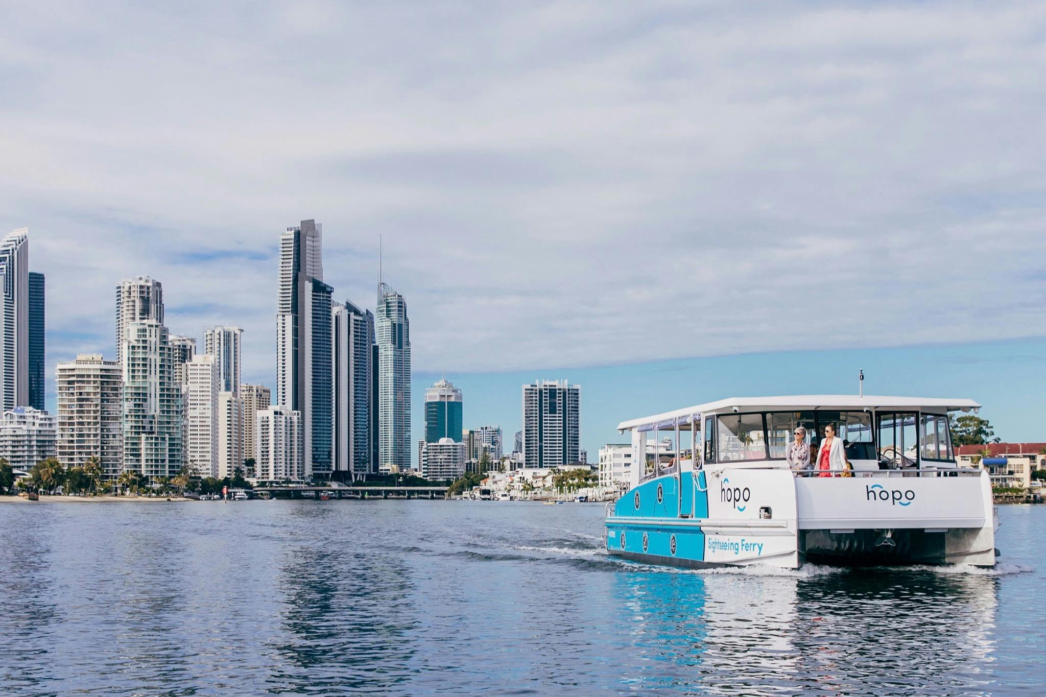 Голубой экскурсионный паром на реке с людьми на борту и городской пейзаж с высокими зданиями на заднем плане.